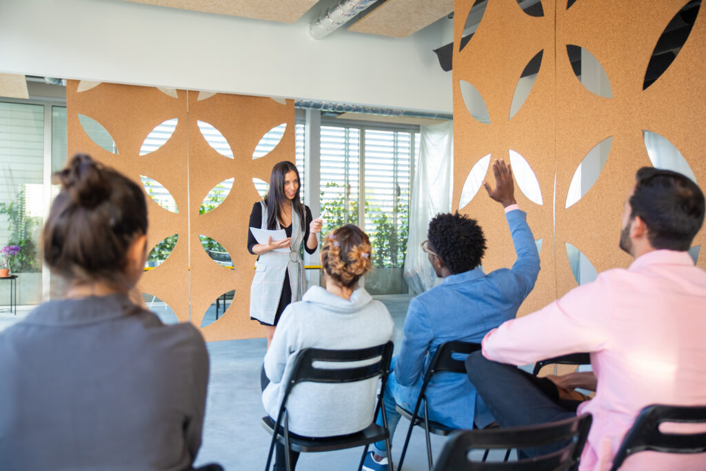 A young woman is giving a presentation to an audience. Someone raises their arm to ask a question.